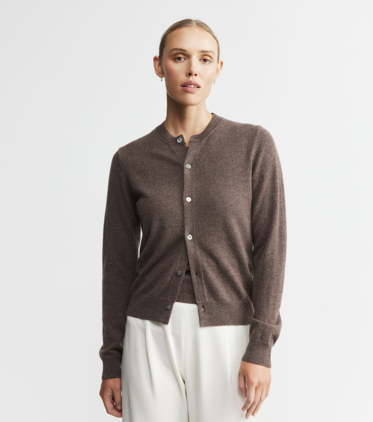 Woman wearing a brown cardigan over a black top with white pants on a light gray background